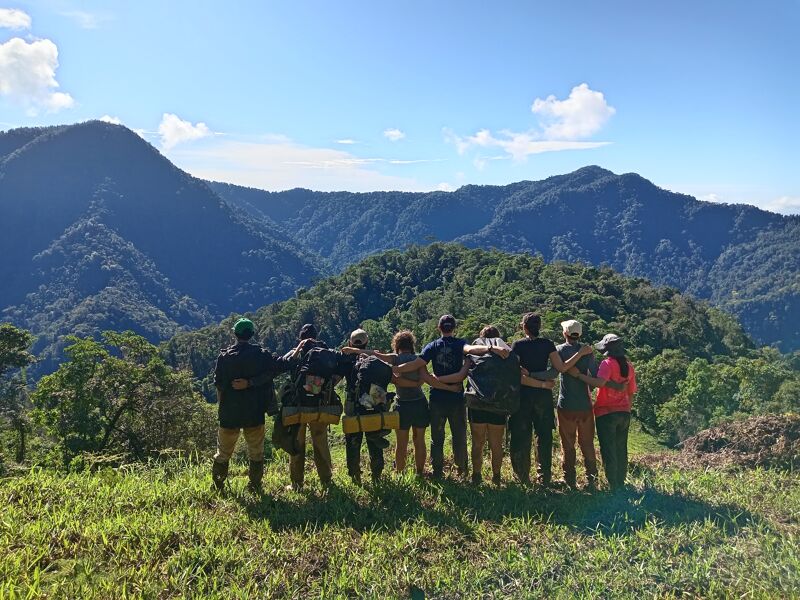 A group of people stands on a grassy hilltop, their backs to the camera, gazing out at a vast, mountainous landscape. The sky is blue with scattered clouds. The people appear to be a team or group, possibly hikers or conservationists, enjoying the scenic view after a trek. The lush greenery suggests a tropical or temperate climate.